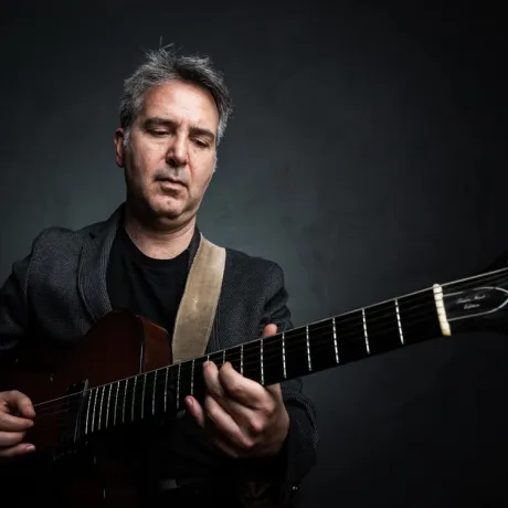 Ein Mann mit kurzen grauen Haaren im grauen Sacko spielt Gitarre und schaut dabei den Gitarrenhals an.