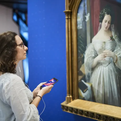 Young woman stands with smartphone and headphones in front of the portrait of Albertine Heine dressed as the bride of August Theodor Kaselowsky