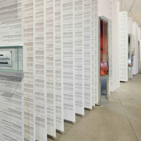 Long white paper banners with text hang from the ceiling interrupted by showcases, in the background stands a blonde woman