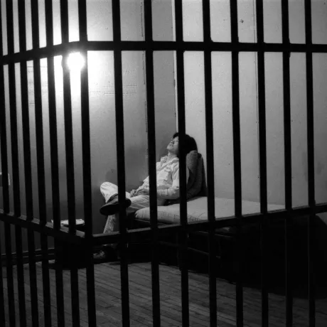 Schwarz-Weiß-Foto eines Menschen hinter Gitterstäben, der auf einer Pritsche sitzt.