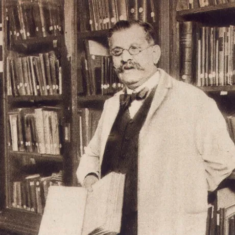 Schwarz-weiß-Foto: Magnus Hirschfeld mit Brille, Schnurbart und weißem Kittel. Er steht vor einem Buchregal und schaut freundlich in die Kamera. 