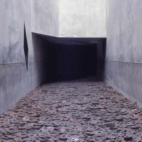 Ein Korridor mit kahlen Betonw&auml;nden, der sich in der Dunkelheit verliert. Der Boden ist &uuml;bers&auml;t mit dicken Metallscheiben, in die Gesichter mit aufgerissenen M&uuml;ndern ges&auml;gt wurden.