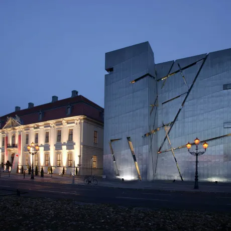 The front of the Old Building and the Libeskind Building are lit up under the darkening night sky