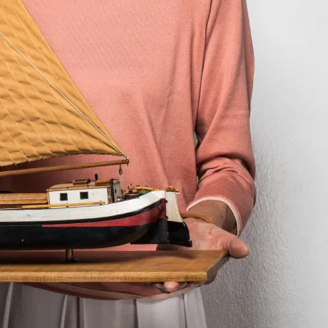 Hände halten ein Holzbrett, auf dem ein Modellschiff steht.