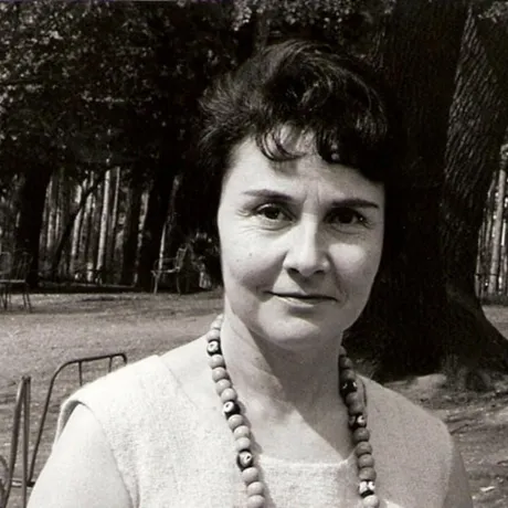 Schwarz-Wei&szlig;-Foto einer mittelalten Frau mit kurzen dunklen Haaren und auff&auml;lliger Kette um den Hals, die freundlich-skeptisch in die Kamera schaut, im Hintergrund B&auml;ume.