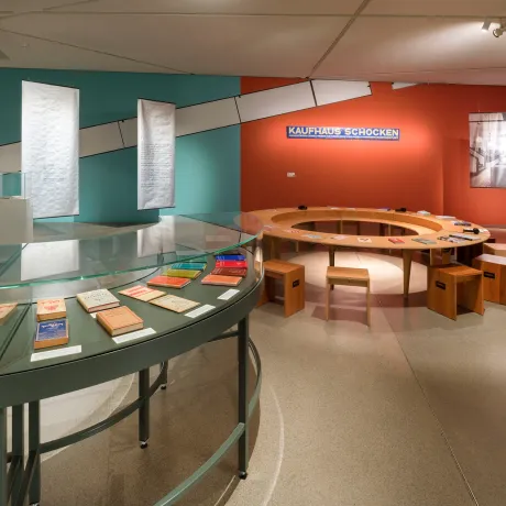 View into an exhibition room, in the foreground a round green display case with books, in the background a round wooden table with chairs, behind it an orange-painted wall with a sign for the Schocken department store.