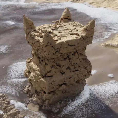 Quadratisch in die Höhe ragender Sandturm am Strand mit vier spitzen Zinnen an den oberen Ecken, von Meerwasser umspült.