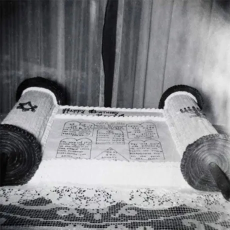 Black-and-white photo of a cake shaped like a Torah scroll on a table with a lace tablecloth