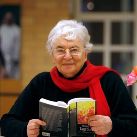 Frau mit aufgeschlagenem Buch in den Händen schaut freundlich in die Kamera.