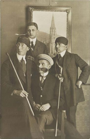 Black-and-white photograph: four student fraternity members, two of them with headbands, in front of a photo of the Freiburg 