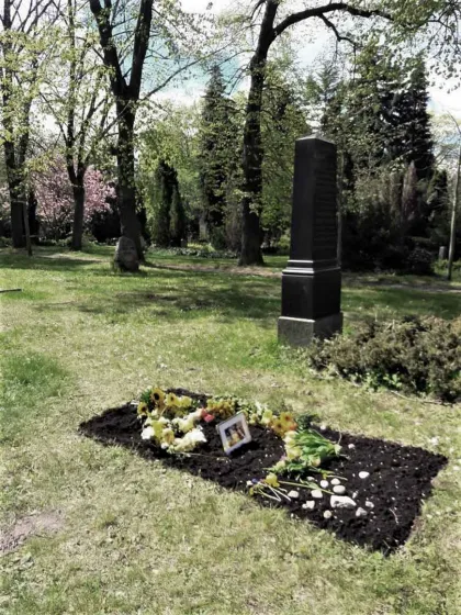 Ein frisches Grab, auf dem Blumen, kleine Steine liegen und ein gerahmtes Foto steht.
