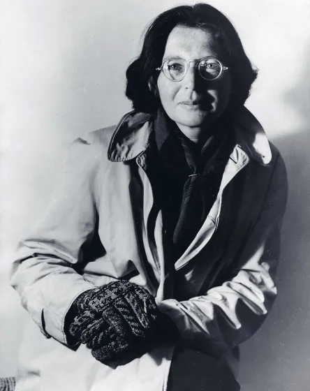 Schwarz-weiß-Fotografie einer Frau, die einen hellen Mantel und schwarze Handschuhe trägt und sich damit auf ihrem aufgestellten Knie abstützt.