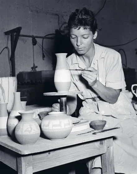 Eine schwarz-weiß-Fotografie zeigt eine junge Frau an einem Tisch mit Keramikgut sitzend. Sie bemalt eine Form, die vor ihr auf einem kleinen Podest steht.