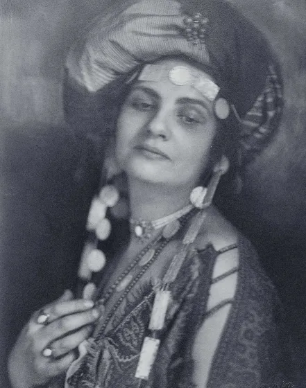 Die sepaifarbene Fotografie zeigt eine Frau mit Turban und einer Tunika und auffälligem Schmuck. Ihr Oberkörper und Gesicht sind zur Kamera gewendet aber ihr Blick geht Richtung Boden.