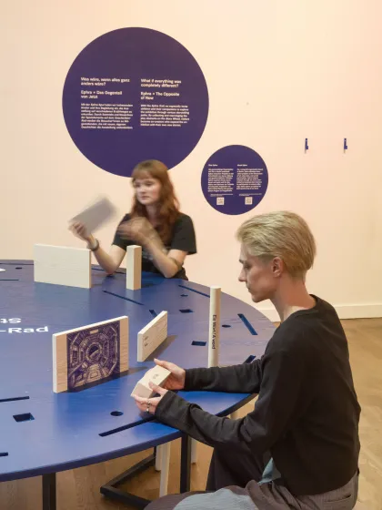 Zwei Menschen sitzen einem runden Tisch in einem Ausstellungsraum und schauen sich Objekte aus Holz an. 