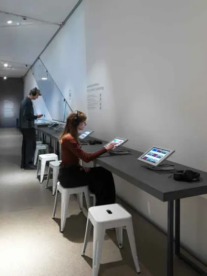 Das Bild zeigt einen Ausstellungsraum. Auf der rechten Seite an der Wand befinden sich schwarze Tische, auf denen Tablets angebracht sind. Vor den Tischen steht ein Mensch und auf einem der weißen Hocker sitzt eine Person.
