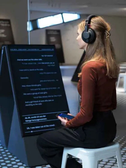 Das Bild zeigt eine Frau mit Kopfhörern, die auf einem weißen Hocker sitzt. In der Hand hält sie ein Gerät. Neben ihr steht ein Aufsteller mit Text.