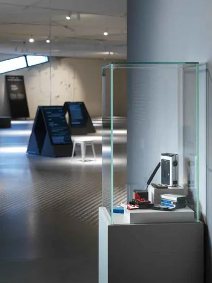 Das Bild zeigt einen Ausstellungsraum, in dem im Hintergrund weiße Hocker und Aufsteller mit Textverteilt sind. Im Vordergrund rechts an der Wand steht eine Vitrine, in der Magnetkassetten ausgestellt sind.