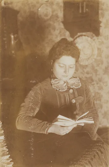  Sepia-colored portrait of a woman holding a book in her hand and appearing to be reading.