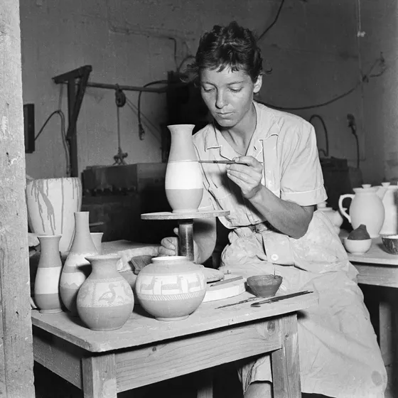  A black and white photograph shows a young woman sitting at a table with ceramics. She is painting a mold that stands on a small pedestal in front of her.