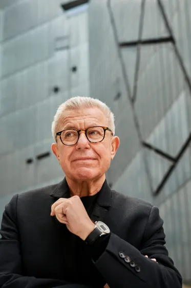 Portait Foto von Daniel Libeskind. Im Hintergrund ist der Libeskind Bau des J&uuml;dischen Museum Berlin unscharf zu sehen.