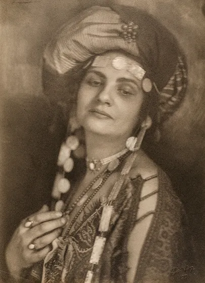  The sepia-colored photograph shows a woman wearing a turban and a tunic with eye-catching jewelry. Her upper body and face are turned towards the camera but her gaze is directed towards the ground.