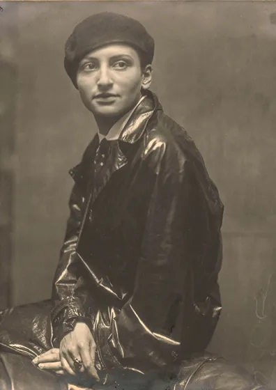  Sepia-colored portrait of a woman whose hair disappears completely under a small cap. She looks past the camera, her upper body turned sideways. She is wearing a leather jacket.