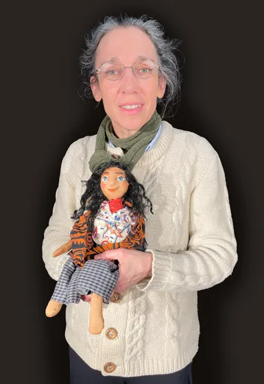 The picture shows the body portrait of a woman up to her hips. She is wearing glasses and her hair is gray. She is holding a doll in her hand.