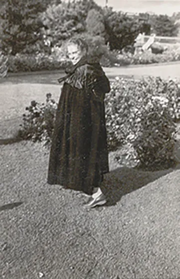 The black and white photo shows a smiling woman in a landscape setting wearing a coat.