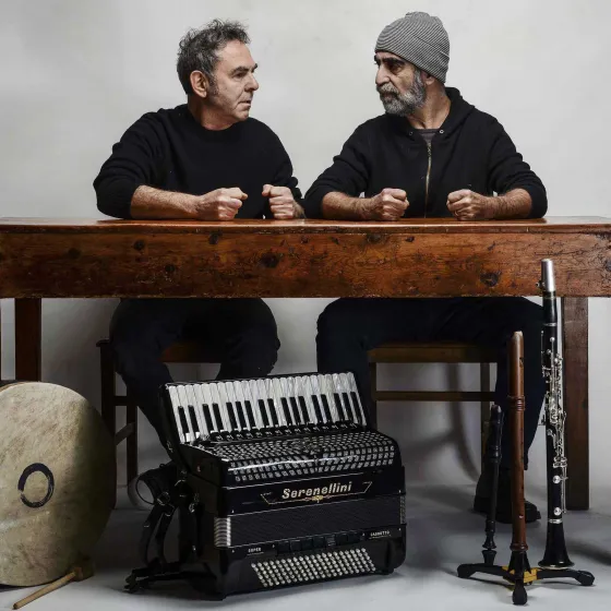 Two men are sitting next to each other at a wooden table, looking at each other, their four fists clenched on the tabletop. In front of the table are five instruments: a drum, an accordion, two flutes and a clarinet.
