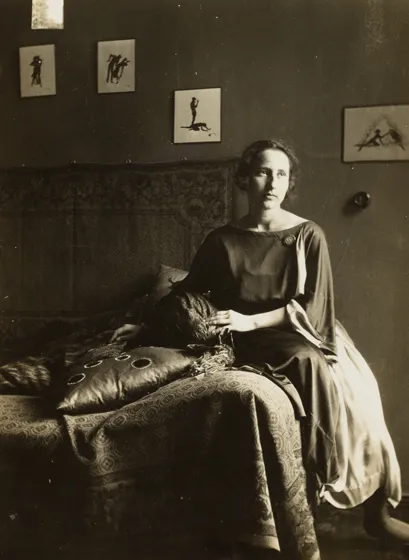  Sepia-colored portrait of a woman with chin-length hair. She is looking past the camera, wearing a dress and sitting on a sofa or bed.