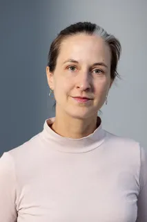 Eine Frau ist mit Gesicht und Oberkörper abgebildet, sie trägt einen rosafarbenen Pullover und blick direkt in die Kamera.