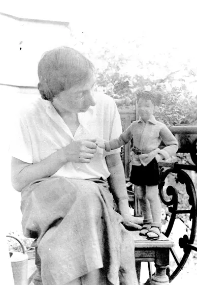 Black and white photo of a woman standing next to a doll on the right, holding its hand and looking at it.