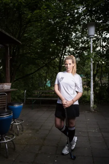 Eine Frau mit blonden Haaren blickt in die Kamera. Sie tr&auml;gt kniehohe, schwarze Str&uuml;mpfe, Shorts und ein Trikot. Mit den H&auml;nden st&uuml;tzt sich sich auf einem Hockeyschl&auml;ger ab.