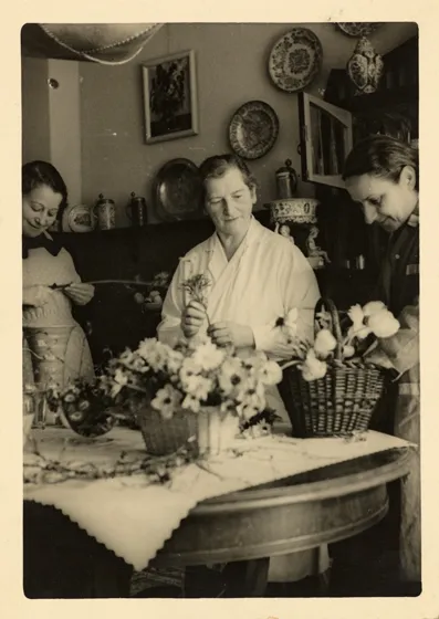  Sepia-colored photograph showing three women arranging flower arrangements in wicker baskets.