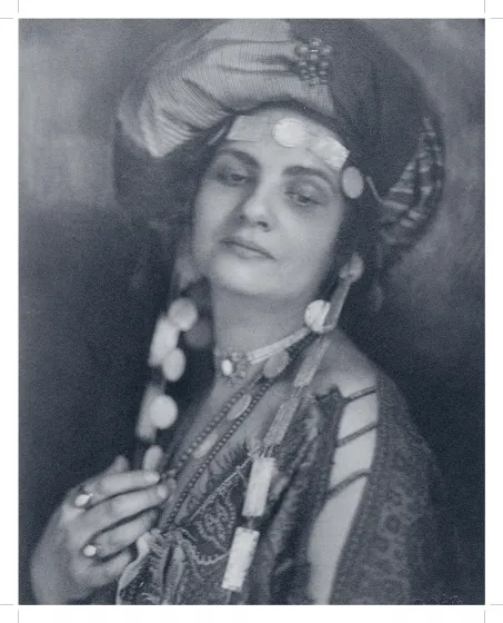 Die sepaifarbene Fotografie zeigt eine Frau mit Turban und einer Tunika und auffälligem Schmuck. Ihr Oberkörper und Gesicht sind zur Kamera gewendet aber ihr Blick geht Richtung Boden.