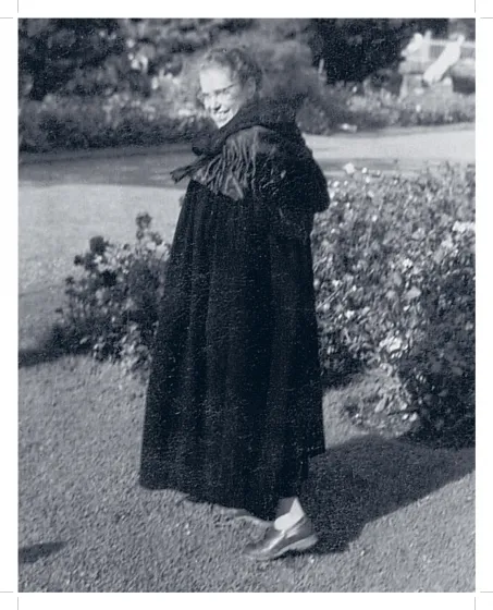 Schwarz-Weiß-Foto einer Frau in einem Garten. Sie trägt einen Mantel, eine Brille und lächselt.
