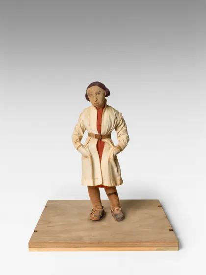 Das Bild zeigt eine Puppe, die auf einem Holzbrett steht. Die Puppe ist eine Frau mit kurzen Haaren und einem weißen Mantel.