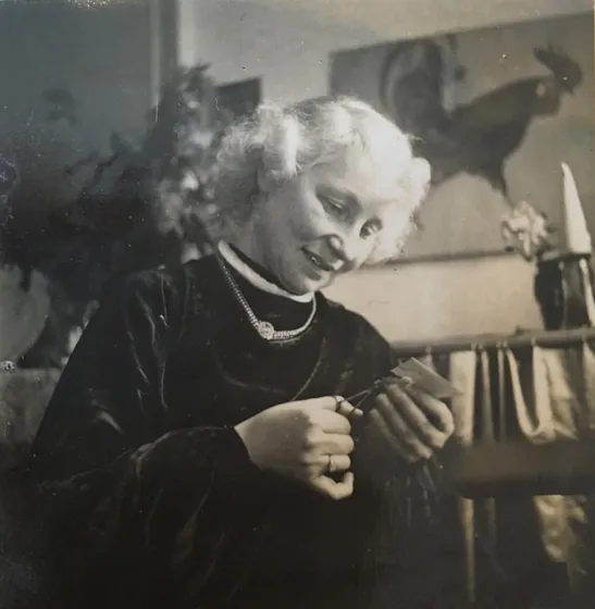  Black and white photograph of a woman looking at a piece of paper she is cutting. There is a shelf behind her and a picture of a cockerel hanging on the wall.