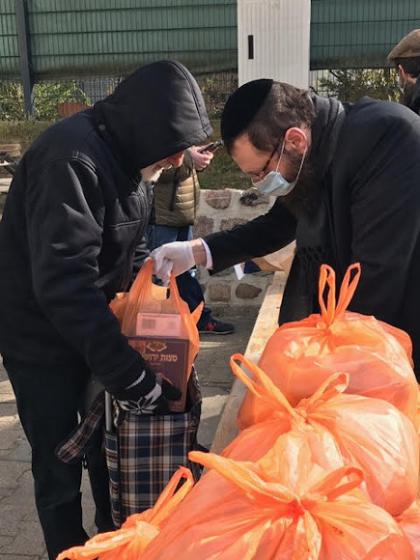 Ein Mann mit Kippa, Mundschutz und Plastikhandschuhen gibt eine orangene Plastiktüte an einen Mann in einer schwarzen Jacke, der einen Stoffbeutel aufhält. Vor den beiden Männer liegen weitere orangene, gefüllte Plastiktüten.