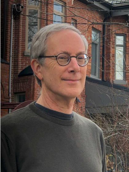 Portrait of a gray-haired man with glasses in front of the brick facade of a house.