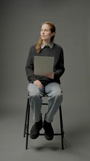 Eine Frau mit langen roten Haaren und dunklem Pullover sitzt auf einem hohen Hocker und h&auml;lt ein Din A4-Skript in der Hand, ihr Blick ist zur Seite gerichtet.