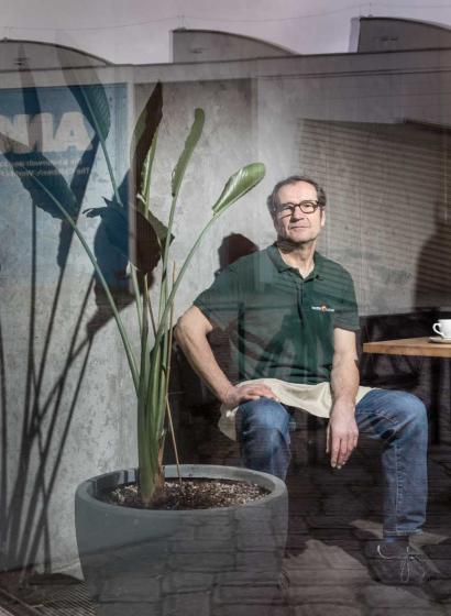 A man in a green shirt with the inscription Beumer&Lutum is sitting at a table. Next to him is a large houseplant. 