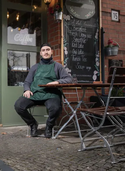 A young man in a dark green apron is sitting at a garden table in front of a café. There is a menu in the background.