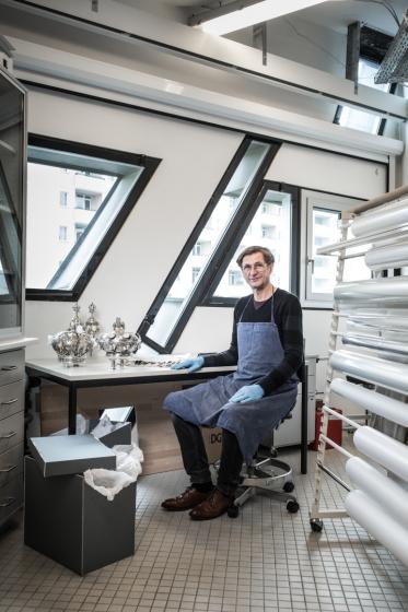 Ein Mann sitzt in einem Dachgeschoss-Atelier an einem Arbeitstisch, vor ihm stehen silberne Objekte, verwendet als Judaica