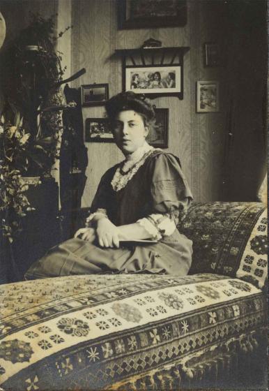 Black and white photo of a young woman. She sits on a divan with oriental-looking pattern and looks into the camera.