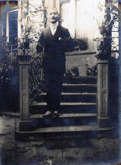 Young man on a staircase in front of a house. He looks mischievous and has both arms bent, it is not possible to tell exactly whether or not he has placed his hands in his jacket pockets.