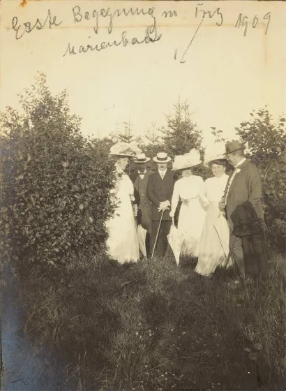Drei Herren in Anzug mit Hut und Stock und drei Damen in weißen Kleidern mit großen Hüten und Sonnenschirmen auf einer Lichtung. Oben auf dem Foto steht in Handschrift: „Erste Begegnung m Fritz Marienbad 1909“, ein Pfeil weist auf Fritz.