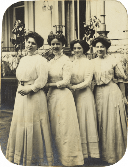 Black and white photo showing four young ladies standing in a row diagonally behind each other in three-quarter profile in front of a house. All are wearing white, high-necked dresses and updos.