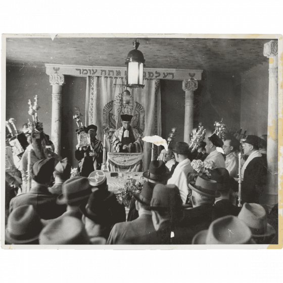 Schwarz-weiß Fotografie einer Synagoge, in der Mitte ein Rabbi, um ihn herum Männer mit Judaica 